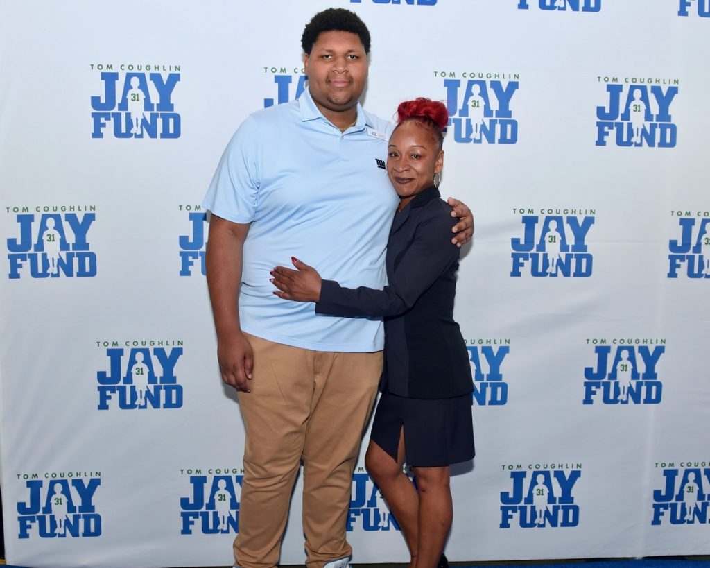A young man and woman smiling and embracing at a charity event. The backdrop features the "Tom Coughlin Jay Fund" logo.